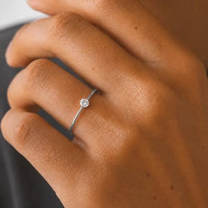 Könnte beinhalten: Ein dünner silberfarbener Metallring mit einem einzelnen kleinen, runden, klaren facettierten Stein in einer einfachen Krappenfassung. Die Ringschiene ist glatt und schmal und umschließt den Finger. Der Text SM ESSENCE steht unten im Bild.