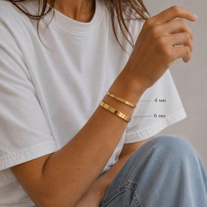 May include: Gold-colored bracelets, 4 mm and 6 mm wide, are displayed on a person's wrist. The bracelets are simple, flat bands. The person is wearing a white t-shirt and blue jeans. The brand name "SMESSENCE" is visible at the bottom of the image.