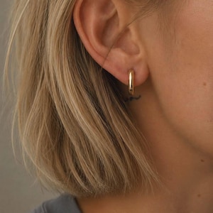 May include: A small, polished gold hoop earring with a smooth, rounded surface. The earring is circular in shape and features a simple, continuous design. The text SM ESSENCE is printed in white at the bottom of the image frame.