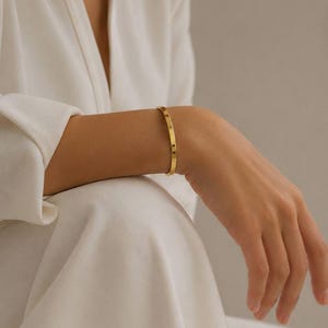 May include: A gold-colored bangle bracelet with a minimalist design. The bracelet is worn on a wrist, and the image includes the text "SM ESSENCE". The bracelet appears to be made of metal and has a smooth, polished surface.