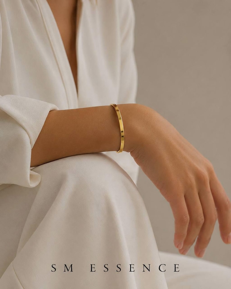May include: A gold-colored bangle bracelet with a minimalist design. The bracelet is worn on a wrist, and the image includes the text "SM ESSENCE". The bracelet appears to be made of metal and has a smooth, polished surface.