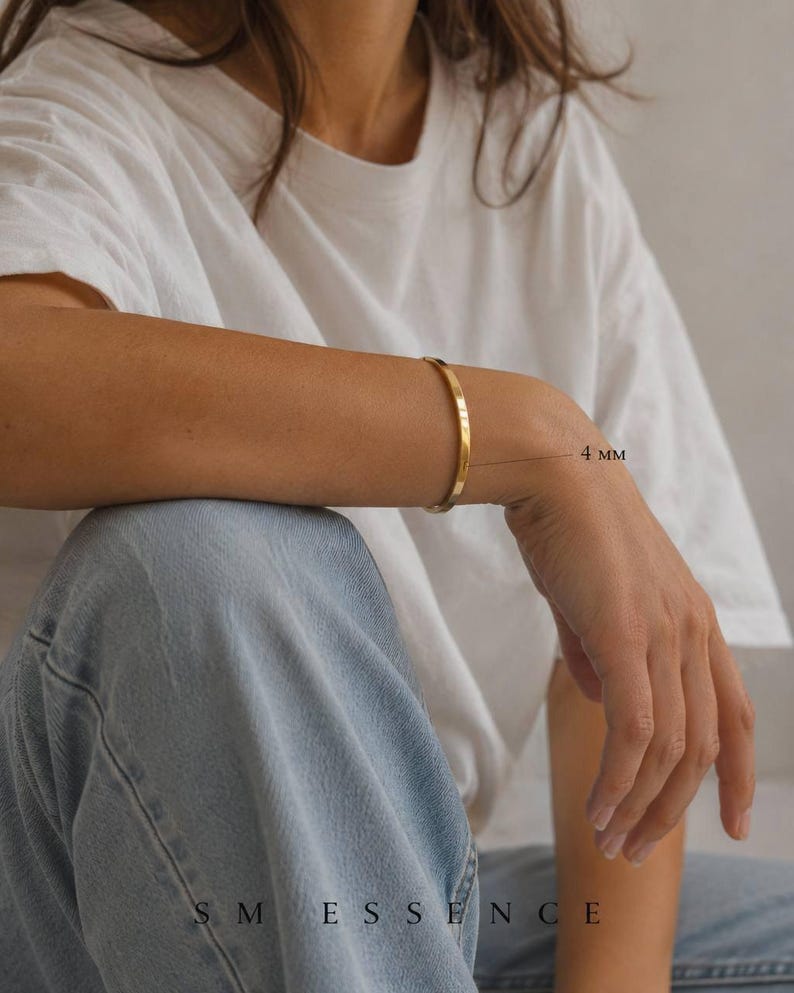 May include: A gold-colored, 4 mm wide, minimalist bracelet is worn on an arm. The person is wearing a white t-shirt and blue jeans. The brand name "SM ESSENCE" is visible at the bottom of the image.