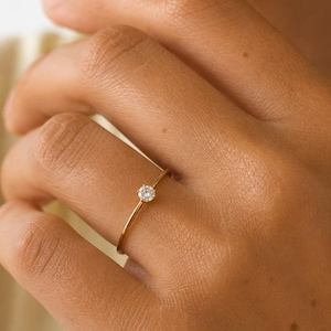 Puede incluir: Un anillo fino de oro con una pequeña piedra preciosa redonda y transparente en un engaste sencillo de garras. El aro es liso y pulido, con un diseño sencillo. El texto SM ESSENCE aparece impreso en negro en la parte inferior de la imagen.