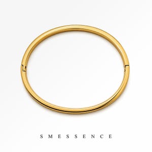 May include: A gold-colored bangle bracelet with a hinged clasp. The bracelet is a simple, circular design with a smooth, polished finish. The word "SMESSENCE" is printed in black, sans-serif font below the bracelet.