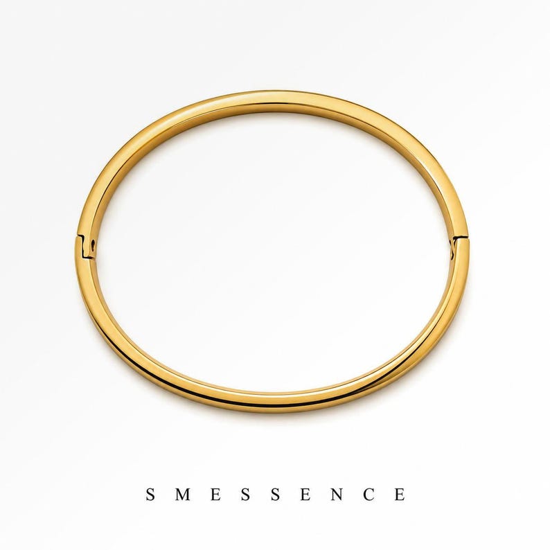 May include: A gold-colored bangle bracelet with a hinged clasp. The bracelet is a simple, circular design with a smooth, polished finish. The word "SMESSENCE" is printed in black, sans-serif font below the bracelet.