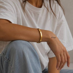 May include: A gold-colored bangle bracelet, 6 mm wide, is worn on a person's wrist. The bracelet has a smooth, polished surface. The person is wearing a white t-shirt and blue jeans. The brand name "SM ESSENCE" is visible.