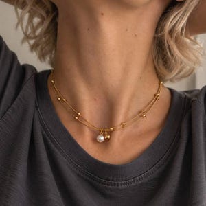 May include: A double-strand gold chain necklace featuring small gold beads along the chains and two central charms, one white pearl and one smooth gold sphere. The text SM ESSENCE is printed in white at the bottom of the image.