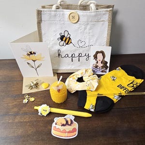 May include: A gift set featuring a white burlap tote bag with a bee graphic and the word happy, a bee-themed greeting card, yellow socks with black accents, a honeycomb-shaped candle, a yellow pen, a scrunchie, a bee sticker, and charms.