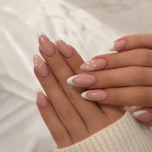 Puede incluir: Uñas almendradas con manicura francesa. Las uñas tienen una base nude con puntas blancas y pequeños diseños florales blancos en algunas. Las uñas están bien cuidadas y tienen un acabado brillante.