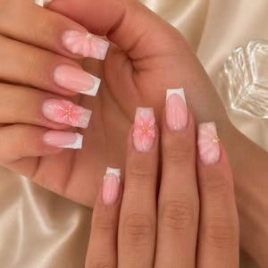 Puede incluir: Una mano con manicura con uñas acrílicas rosas y blancas. Algunas uñas tienen un diseño floral 3D con detalles dorados. Las uñas son de forma cuadrada con un diseño de punta francesa. El fondo es una tela satinada.