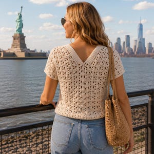 May include: A short-sleeved cream-colored crochet top with a V-neckline and an intricate openwork pattern, paired with light blue denim jeans and a tan woven straw tote bag carried over the shoulder, showcasing a textured knit construction.