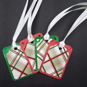 May include: Four rectangular gift tags with white ribbon. The tags have a red and green glitter border and a plaid pattern in red, green, and white. The tags are likely for Christmas or holiday gifts.