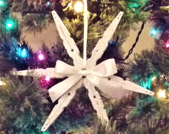 May include: A white, glittery snowflake Christmas ornament with a white satin bow. The snowflake is attached to a green Christmas tree with colorful lights in the background. A festive holiday decoration.
