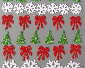 May include: Sheet of Christmas-themed glitter stickers. The stickers include green glitter Christmas trees, white glitter snowflakes, and red glitter bows. The stickers are arranged in rows on a gray background.
