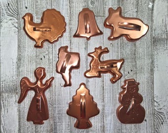 May include: A collection of copper-colored cookie cutters in various shapes, including a turkey, bell, rabbit, angel, pilgrim, reindeer, Christmas tree, and snowman. The cutters are arranged on a light wood-grain surface.