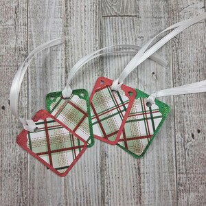 May include: Four decorative gift tags with a red and green plaid pattern and white ribbon. The tags have a red or green glitter border and are diamond-shaped with rounded corners. The background is a light wood grain.