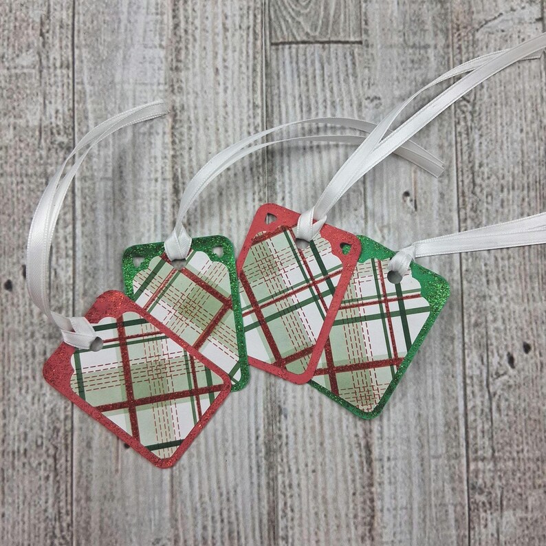 May include: Four decorative gift tags with a red and green plaid pattern and white ribbon. The tags have a red or green glitter border and are diamond-shaped with rounded corners. The background is a light wood grain.