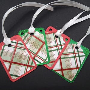 May include: Four diamond-shaped gift tags with white ribbon. The tags have a plaid pattern in red, green, and white. The edges are glittered in red and green. Each tag has a heart-shaped cutout.