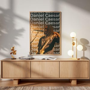 May include: A rectangular framed wall poster featuring a photograph of Daniel Caesar performing with a guitar and microphone. The background of the poster displays the name Daniel Caesar repeated in a grid pattern with a dark gray and orange color scheme.