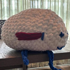 May include: A large, plush, white crocheted creature with red and blue accents. The creature has a rounded body, a red wing-like detail, blue eyes, and blue, rope-like legs. The handmade item is sitting on a wooden surface.