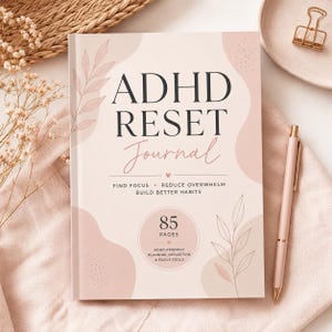 Peut inclure: Un journal rose pâle avec le titre ADHD RESET Journal imprimé en noir. La couverture présente des dessins au trait de feuilles, des formes abstraites et le texte Find Focus, Reduce Overwhelm, Build Better Habits. Un badge indique 85 pages.