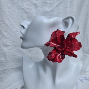 May include: A large, glossy red flower-shaped earring featuring layered, organic petals with a smooth, reflective finish. The earring is displayed on a white mannequin bust, highlighting the bold, sculptural design and vibrant red color of the piece.