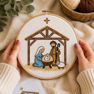 Può includere: Un telaio da ricamo circolare in legno contenente un motivo a punto croce di una scena della natività. Il disegno presenta una stalla marrone, una figura vestita di blu, una figura vestita di marrone con un bastone, un bambino e una croce.