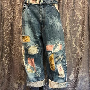 May include: Blue acid-wash denim capri pants featuring a button and zipper closure, belt loops, and multiple rectangular fabric patches in various floral and patterned prints. The hem of each pant leg is cuffed with a grey and white floral fabric.