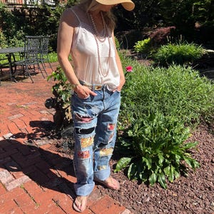 May include: A light pink satin camisole with lace trim, a wide-brimmed straw hat, and long layered pearl necklaces paired with distressed blue denim jeans featuring various colorful fabric patches and a metallic belt with a rectangular buckle.