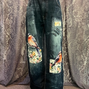 May include: Dark blue denim jeans featuring distressed areas, frayed hems, and decorative patches. The patches display colorful bird illustrations and floral patterns. The waistband includes a small floral fabric accent and a single button.