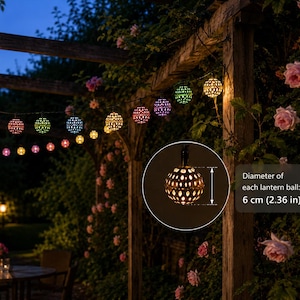 Puede incluir: Una cadena de luces LED multicolores con pantallas esféricas de metal perforado. Cada bola de linterna tiene un diámetro de 6 cm, como indica el texto superpuesto. Las luces están suspendidas al aire libre y emiten un brillo cálido.