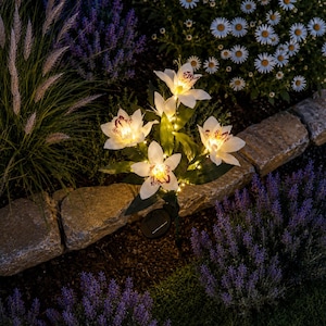 Puede incluir: Una lámpara solar de jardín con forma de estaca que presenta cuatro flores blancas tipo lirio con centros moteados en rojo oscuro. Los pétalos están iluminados desde el interior por pequeñas luces LED cálidas y la base incluye un panel solar negro.