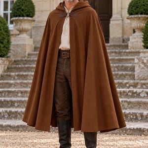 May include: A floor-length brown hooded cloak featuring a dark brown leather strap closure with a metal ring. The cloak is worn over a light beige long-sleeved shirt, dark brown trousers with a brown leather belt, and tall black leather boots.