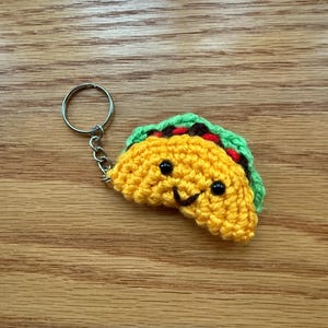 May include: A handmade crochet taco keychain with a smiling face. The taco is yellow with green and brown accents, and black bead eyes. The keychain has a silver metal ring and chain.