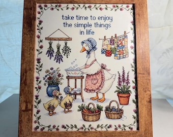 May include: A framed cross-stitch embroidery featuring a goose wearing a bonnet and apron, washing clothes in a basin. The text reads, "take time to enjoy the simple things in life." The scene includes two ducklings, hanging quilts, and baskets.