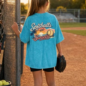 Może przedstawiać: Turkusowa koszulka z napisem "Softballs and Sunsets" z tyłu. Grafika zawiera piłkę do baseballu, palmy i zach&oacute;d słońca. Osoba ma na sobie czarne szorty i skarpetki i trzyma rękawicę do baseballu.