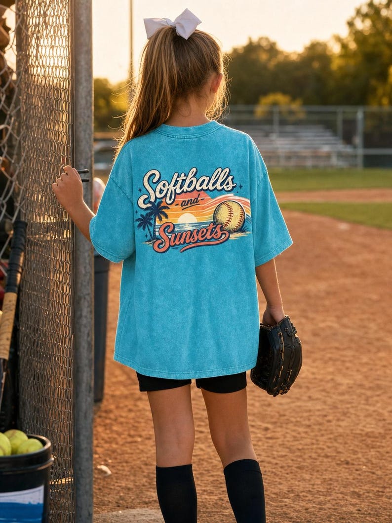 Może przedstawiać: Turkusowa koszulka z napisem "Softballs and Sunsets" z tyłu. Grafika zawiera piłkę do baseballu, palmy i zach&oacute;d słońca. Osoba ma na sobie czarne szorty i skarpetki i trzyma rękawicę do baseballu.
