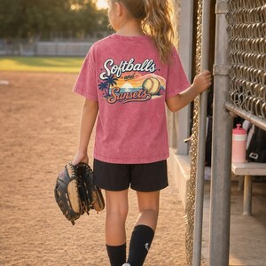 Może przedstawiać: Osoba nosi czerwoną koszulkę z napisem "Softballs and Sunsets" z tyłu, wraz z piłką do softballu i palmami. Mają na sobie czarne szorty, białe trampki i czarne skarpetki. Trzymają rękawicę do baseballu.