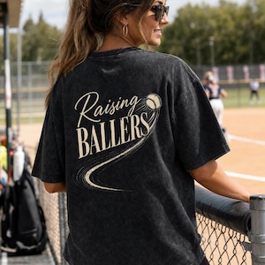 Op de afbeelding: Donkergrijs t-shirt met de tekst "Raising Ballers" en een honkbalafbeelding. Het shirt heeft een vintage, gewassen look en een ronde hals. Het ontwerp is cr&egrave;mekleurig.