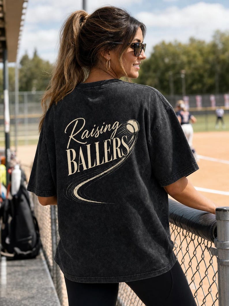 Op de afbeelding: Donkergrijs t-shirt met de tekst "Raising Ballers" en een honkbalafbeelding. Het shirt heeft een vintage, gewassen look en een ronde hals. Het ontwerp is cr&egrave;mekleurig.