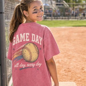 May include: A dusty rose t-shirt with the words "GAME DAY" above a baseball graphic. Below the baseball, the text reads "all day, every day". The shirt is worn by a person on a baseball field.