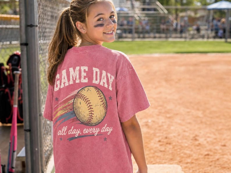May include: A dusty rose t-shirt with the words "GAME DAY" above a baseball graphic. Below the baseball, the text reads "all day, every day". The shirt is worn by a person on a baseball field.