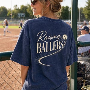 Op de afbeelding: Marineblauw T-shirt met de tekst "Raising Ballers" in een cursief lettertype en een honkbalafbeelding. Het shirt heeft een vintage wassing en korte mouwen. De afbeelding toont de achterkant van het shirt.