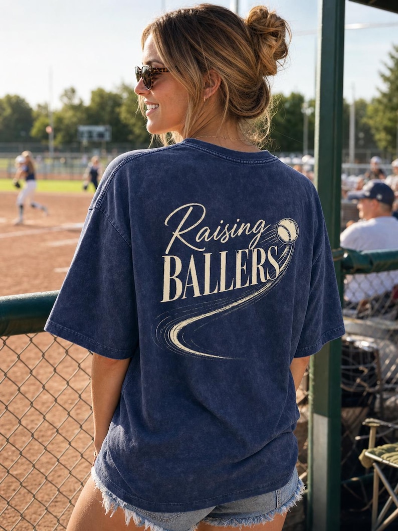 Op de afbeelding: Marineblauw T-shirt met de tekst "Raising Ballers" in een cursief lettertype en een honkbalafbeelding. Het shirt heeft een vintage wassing en korte mouwen. De afbeelding toont de achterkant van het shirt.