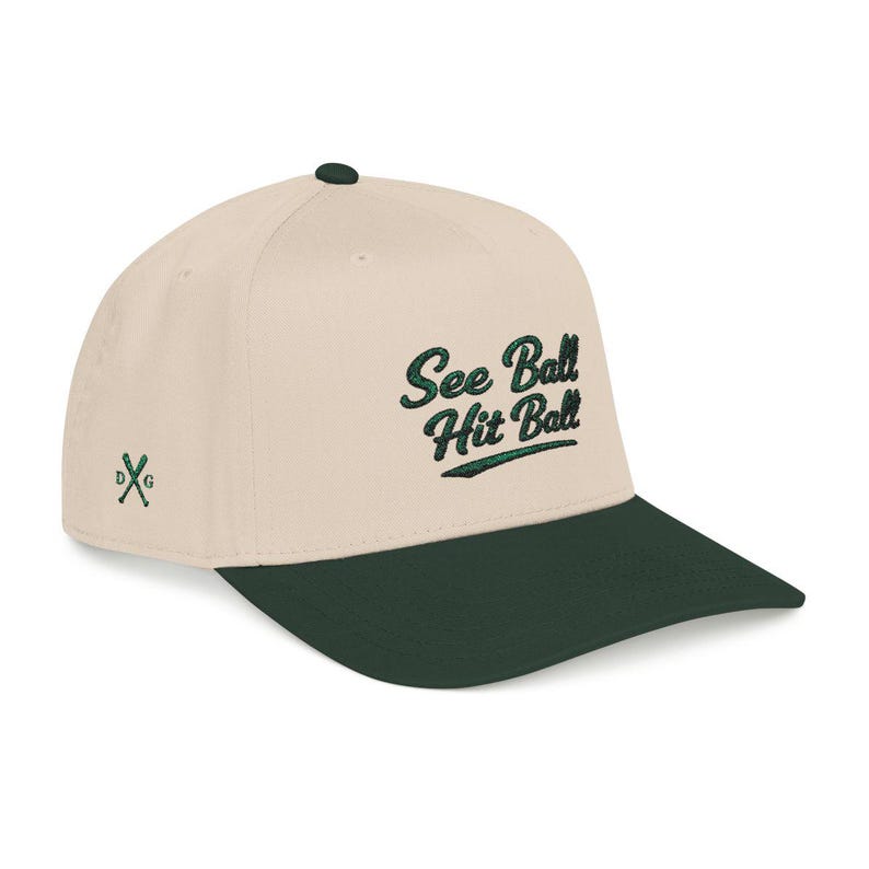 Puede incluir: Gorra de b&eacute;isbol con corona color crema y visera verde oscuro. La gorra presenta el texto bordado "See Ball Hit Ball" en letras verdes. Un peque&ntilde;o logotipo de palos de golf cruzados est&aacute; en el lateral.