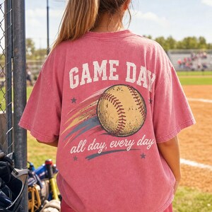 May include: A pink t-shirt featuring the words "GAME DAY" and a baseball graphic. The text "all day, every day" is below the baseball. The shirt is worn at a baseball field.