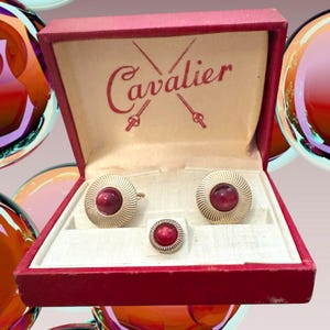 May include: A set of two round gold-tone cufflinks and one matching tie tack, each featuring a deep red cabochon center and a textured sunburst border. The items are presented in a red box with the word Cavalier printed in red script.