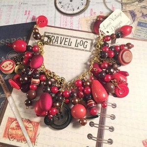 May include: A charm bracelet featuring a gold-tone chain adorned with an assortment of red and brown beads, buttons, and spherical charms. A small white tag attached to the bracelet displays the text Lunas Vintage Designs in black ink.