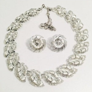 May include: A silver-toned jewelry set featuring a necklace and matching earrings. The necklace has a leaf design, and the earrings are circular with a textured pattern. The set is displayed on a white surface.