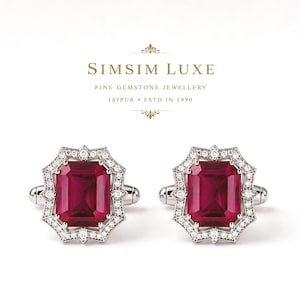 Puede incluir: Un par de gemelos de plata con una gran piedra preciosa rectangular de color rojo intenso en el centro, rodeada por un borde de pequeñas piedras claras y brillantes. El texto SIMSIM LUXE FINE GEMSTONE JEWELLERY JAIPUR ESTD IN 1990 es visible arriba.